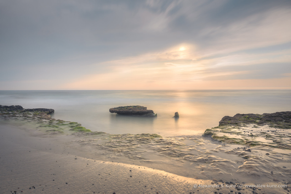 A long exposure seascape photo with coastal rocks in the foreground. The rocks form a small cove and in its center are two rock "islands", one very small and one bigger, with a flat top. The ocean is rendered very smooth with a long exposure time. The sky is mostly cloudy, but the sun peeks through an opening, casting warm light onto the ocean.