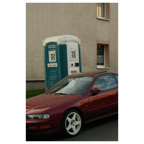 Ein weinroter Sportwagen mit weißen Felgen steht vor einem Dixi-Klo. Auf der Tür steht „toi toi“.  Im Hintergrund ein beige farbenes Haus. 