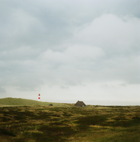 Square format photo of a thatched roof house and a lighthouse sitting in a landscape of dunes.