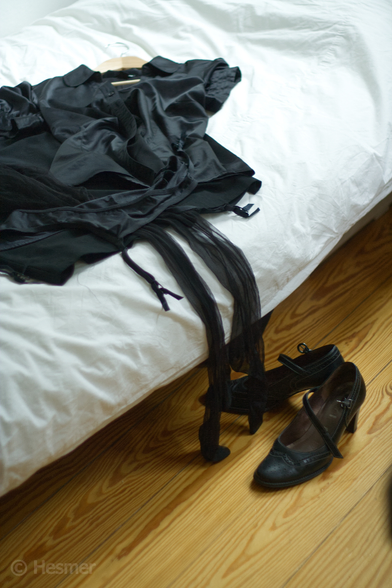 Schwarze Kleidungsstücke liegen auf einem Bett mit weisser Bettwäsche. Nylon-Strümpfe hängen über die Kante, danaben Strapse. Vor dem Bett steht ein Paar hochhackiger schwarzer Schuhe.