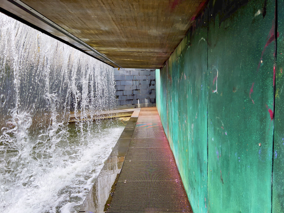 Photo from a sort of an underpass that goes under a waterfall-like water feature outdoors. On the left side of the image is a sheet of water falling into a deeper pool of water (well it's actually a short channel that ultimately leads into the Baltic), and on the right is a strikingly green wall, made from copper that's been oxidised on purpose. Remnants of a purplish red graffiti can be seen on the wall and the walkway. At the end of the walkway is a stone wall, where the walkway takes a sharp turn left