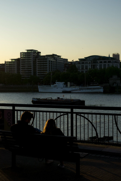 Photo of a couple of people sitting on a bench, facing away from the camera towards a metal railing above a river. A boat is silhouetted against water that reflects a fading sky, past that a white boat stands out against the shadowy far bank on which sits mounds of large buildings. The figures on the bench are mostly dark, though their hair catches the early evening sunlight coming in from the left.