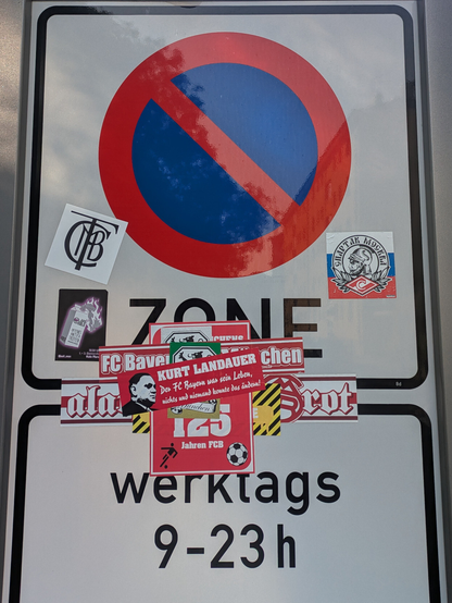 Auf  einem hohen Parkzonen-Schild mit Halteverbot, "ZONE" , "werktags 9 - 23 h" sind zwischen den Texten allerlei Aufkleber auf- und nebeneinander plaziert. In der Mitte dominiert das Rot der Botschaften der Fans des FC Bayern München, rechts Spartak Moskau, MBTC oder FCBM links, überklebtes Grün-Gelb-Schwarz in der Mitte und ein Antifa-Aufkleber vervollständigen die Sammlung. Das Schild spiegelt leicht Bäume und Himmel.