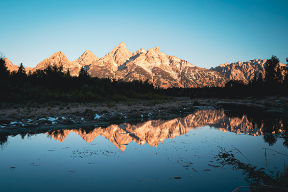 The sun shining directly on sharp peaked mountains mirrored in a still lake with trees lining the shore.
