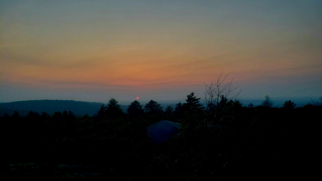 Sunset this evening, June 12th, from Buck Hill! A red disc of the sun sits just above the horizon. Colors of gray, blue and orange paint the sky above the sun.