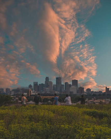 The image depicts a cityscape during sunset. The sky is filled with dramatic, wispy clouds illuminated by the setting sun, casting hues of orange and pink. In the foreground, there are people sitting on a grassy hill, seemingly enjoying the view. The city skyline features several tall buildings, including skyscrapers, suggesting it might be a major urban area. The overall atmosphere is serene and picturesque, capturing a moment of tranquility amidst the urban environment.