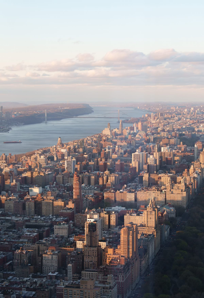 The image depicts a cityscape with a dense cluster of buildings, predominantly mid-rise and high-rise structures. The architecture suggests a bustling urban environment. In the background, a large body of water is visible, with a bridge spanning across it. The skyline on the far side of the water features prominent skyscrapers, indicating a major city. The lighting and shadows suggest it is either early morning or late afternoon. This cityscape resembles that of New York City, with the Hudson River and the George Washington Bridge visible.