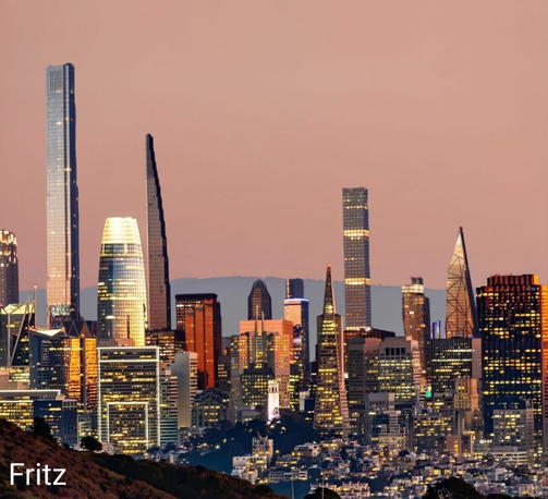 The image depicts a cityscape during dusk or dawn, characterized by a series of illuminated skyscrapers and high-rise buildings. The tallest buildings are prominently lit, casting a warm glow against a pinkish sky. The city appears modern and bustling, with a mix of architectural styles. The name "Fritz" is visible at the bottom left, which might be the photographer's name or a watermark. The background includes faintly visible hills or mountains, adding depth to the urban scene.