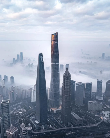 The image depicts a cityscape with a dense cluster of high-rise buildings. Three prominent skyscrapers dominate the foreground, with the tallest one being particularly slender and tapering towards the top. The buildings exhibit modern architectural designs, with glass facades reflecting the light. The city is enveloped in a light mist or fog, giving it a serene and somewhat ethereal atmosphere. The background reveals more buildings, suggesting a bustling urban environment. The city appears to be Shanghai, identifiable by the distinctive skyscrapers.