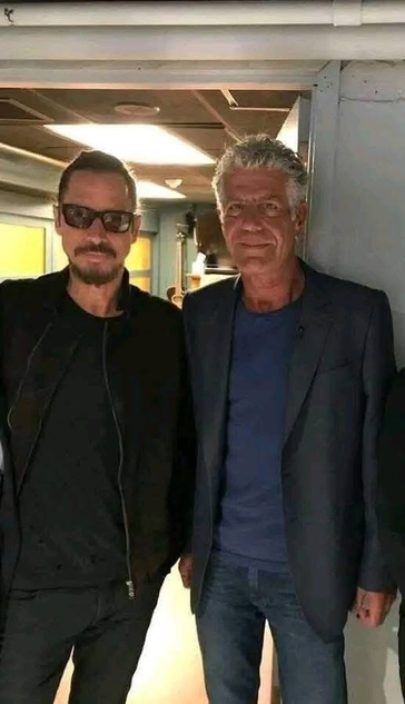 The image features two men standing side by side in what appears to be a backstage or dressing room area. The man on the left is wearing a black jacket, black shirt, and sunglasses, with a mustache. The man on the right is dressed in a dark blazer over a blue shirt and jeans. Both men are smiling and looking directly at the camera. The background includes a white wall, a light fixture, and some indistinct objects. The setting suggests a casual, friendly atmosphere, possibly at an event or gathering.