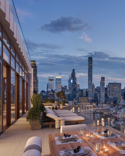 The image depicts a cityscape at dusk, with a skyline featuring numerous high-rise buildings. The sky is painted with hues of blue and purple, and a few clouds are scattered across. In the foreground, there is a rooftop terrace with a dining table set for a meal, complete with plates, glasses, and candles. The terrace also includes a seating area with cushioned benches and a large potted plant. The architecture and skyline suggest that this could be New York City.