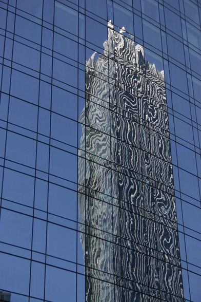 A color photo of a glass skyscraper. One side of the building fills the frame, with another, older, structure appearing reflected and distorted in the glass.