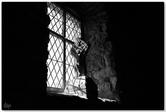 Ein Fenster in einer alten Kirche. In der Fensternische steht ein Stein mit einer menschlichen Figur, die die Arme in Richtung Fensterkreuz hebt. Das Licht wirft Schatten auf den Stein, der Rest des Bildes ist dunkel. 

A window in an old church. In the window recess is a stone with a human figure raising its arms towards the window cross. The light casts shadows on the stone, the rest of the picture is dark.