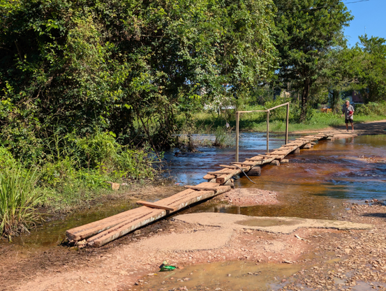 Über eine unbefestigte Straße fließt Wasser. Aus runden Holzstücken und dicken Bohlen wurde ein provisorischer Steg gebaut, der hindurchführt.