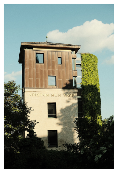 Modernes, mehrstöckiges Gebäude mit einer Fassade aus sandfarbenem Putz und oberen Stockwerken mit bronzefarbener Metallverkleidung. Auf der Fassade ist eine griechische Inschrift zu sehen („ἄριστον μὲν ὕδωρ“ – „Das Beste ist Wasser“). Die rechte Gebäudeseite ist vollständig von grünem Efeu überwachsen. Umgeben ist das Haus von dichtem, sommerlichem Grün unter einem hellblauen Himmel mit vereinzelten Wolken.