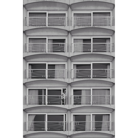 Black and white street photography taken in Belgium. Ten identical windows on a modernist facade create a regular pattern on two columns, with small variations depending on the curtains. An old man wearing a white shirt is standing on the balcony of the fourth window on the left.