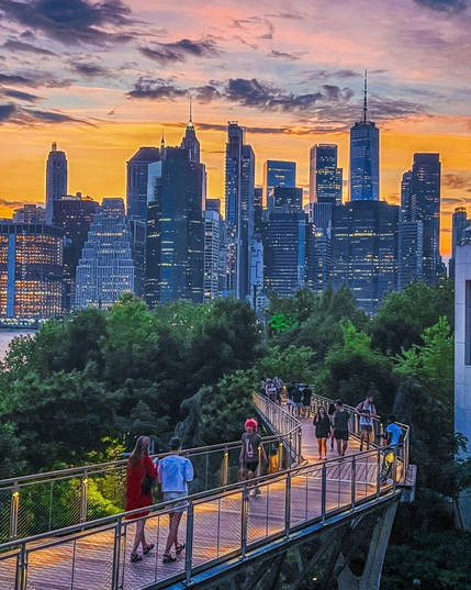 The image depicts a cityscape during sunset. The skyline features numerous high-rise buildings, including a prominent skyscraper with a spire, likely the Empire State Building. The city appears to be New York City, given the recognizable architecture and layout. In the foreground, there is a pedestrian walkway with people strolling and enjoying the view. The sky is painted with hues of orange and pink, adding a dramatic backdrop to the illuminated cityscape.