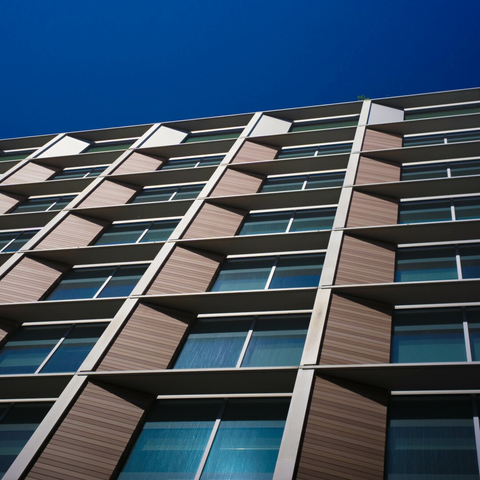 Slanted photo of six floors of identical blue-green windows in harsh sunlight, split vertically into two panes, recessed from a thin square metal grid, a sloping wooden panel on the left of each, more abrupt transitions on the other three sides. Across the top is another storey, similar, but single-paned, and metal-sloped. Above is a slim quadrilateral of clear deep blue sky.
