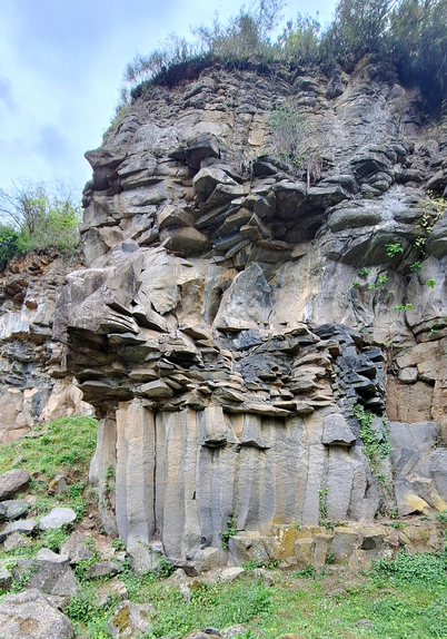 The image depicts a rocky cliff face with distinct geological features. The cliff is composed of large, vertically aligned columns of basalt, characteristic of columnar jointing, which is formed when lava cools and contracts. These columns are arranged in a hexagonal pattern, with some standing upright and others fallen, creating a rugged texture. The rock surface is uneven, with various sizes of rocks and boulders scattered across the base. Vegetation, including grass and small shrubs, is visible at the top of the cliff and on the ground, indicating a natural environment. The sky is partly cloudy, suggesting an overcast day. The overall color palette consists of grays and browns, typical of volcanic rock, with patches of green from the vegetation.

Provided by @altbot, generated privately and locally using Ovis2-8B

🌱 Energy used: 0.191 Wh