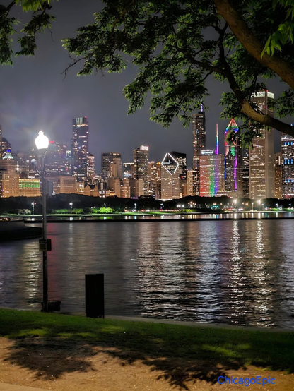 The image depicts a nighttime cityscape with a river in the foreground. The city is illuminated with numerous lights, highlighting various skyscrapers and buildings. A prominent bridge spans the river, connecting both sides of the city. The reflection of the city lights shimmers on the water's surface. Trees and a street lamp are visible in the foreground, adding to the serene ambiance. The city appears to be Chicago, identifiable by the distinctive architecture and the "Chicago" watermark.