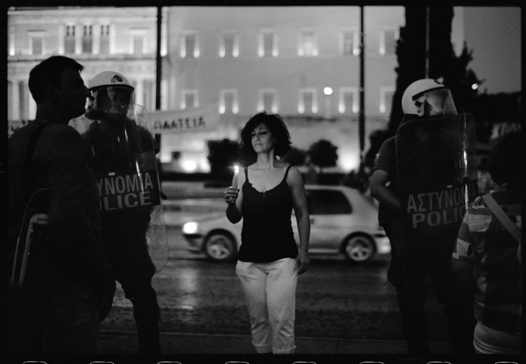 Une femme entourée de policiers tient une bougie. Elle lui éclaire le visage. Il fait nuit. La scène se déroule sur la place Syntagma, à Athènes. On voit en arrière-plan le parlement. 