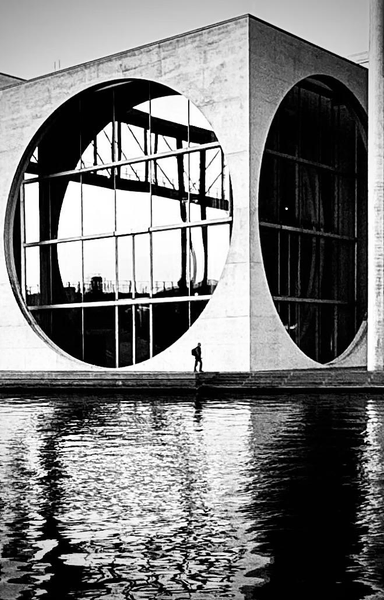 A minimalist black-and-white image of a modern building featuring large circular windows, reflecting a calm body of water. A solitary figure walks along the edge of the water, emphasizing the architectural design.