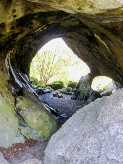 Ein Wanderweg durch eine kleine Höhle. Am anderen Ende ist sind Felsen am Wegrand zu sehen und ein Baum.