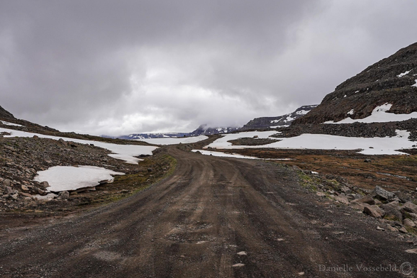 A sandy brownish gravel road, tracks are still visible. The sides of the hills are small rocks, some moss in reen and orange, and then the hills. There are still snow patches on the side of the road and on the hills. The sky is moody. 