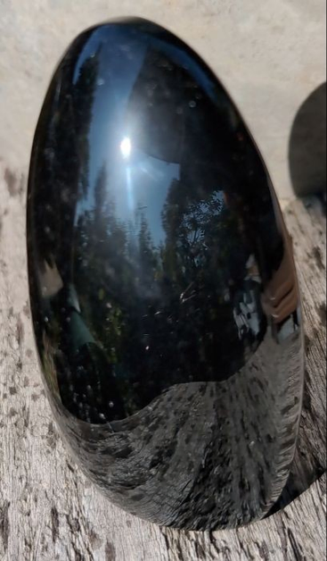 ein als oval halbrund geschliffener, länglicher schwarzer Stein, dessen Oberfläche so glatt ist, dass sie wie ein Spiegel wirkt. Deshalb sind auf dem Stein die Sonne, der blaue Himmel, Bäume, ein Teil einer Solaranlage (rechts oben), ein Teil einer unten aufgeschnittene, grünen Regentonne sowie schön gewölbt das grau verwitterte Holzbrett, auf dem der Stein steht, zu sehen.