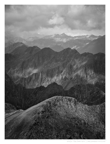Monochrome photograph of a rugged mountain landscape, depicting layers of jagged peaks and ridges receding into the distance, the background progressively turning lighter and mistier. Clouds hover above the peaks, casting dramatic shadows and highlights across the scene. The overall tonal range is rich, from deep shadows in the mountains to bright highlights in the distant clouds.