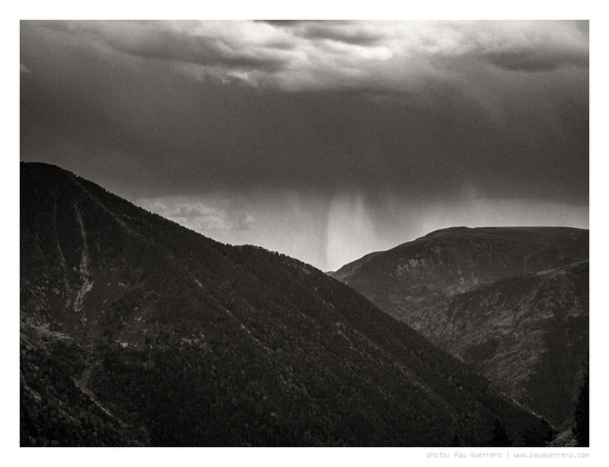 Black and white photograph of a mountainous landscape. Dark clouds overhead and, in between two opposed mountain slopes covered in dark vegetation, a rain shower descending, like a dark Chinese ink smudge. A somber mood commands the scene.
