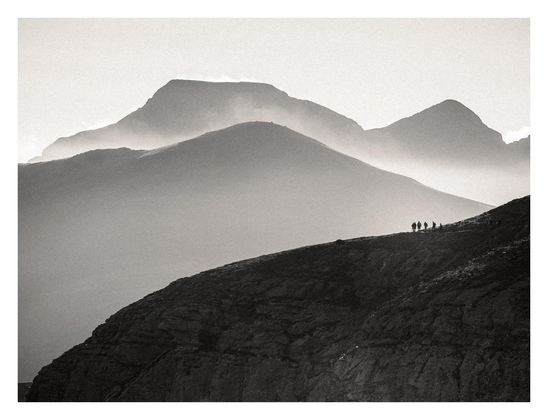 Black and white picture of an early morning mountain landscape, on the foreground we can see a silhouetted group of hikers walking in single file along a narrow track over the edge of a cliff. On the background, layers of hazy mountains receding into the distance mountains enveloped in soft layers of mist.