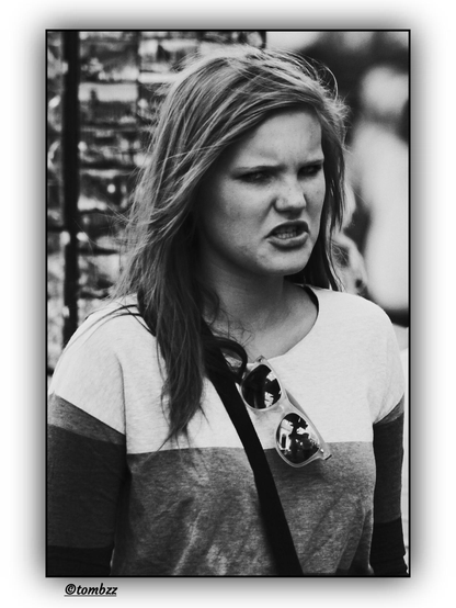 A black-and-white photo shows a young woman standing in front of a souvenir magnet stand. Her face expresses intense emotion — anger, perhaps frustration — which sparks curiosity and gives the image an intriguing character. She’s wearing a long-sleeved top in shades of white, light gray, and dark gray, complementing the monochrome aesthetic of the photograph. A black bag with a strap hangs over her shoulder, and sunglasses are resting on her chest. The background features a stand filled with numerous magnets.