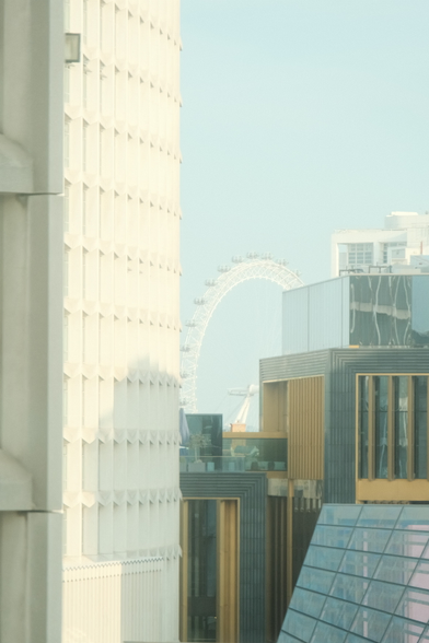 Bright and slightly foggy photo of London eye between buildings.