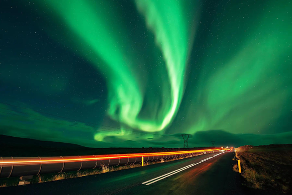 A green wavy aurora winds its way across the nights sky. Below a car leaves a red light trail as it zooms down the road.