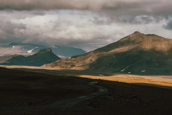 A landscape in Iceland of warm brown mountains, clouds and the hint of a road. In the distance there is snow on a mountain.