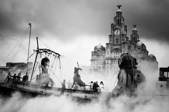A boat sails past the Liverbuilding on a sea of fog. Inside are giant marionettes. A little girl, her dog and her uncle in a steam punk diving suit.