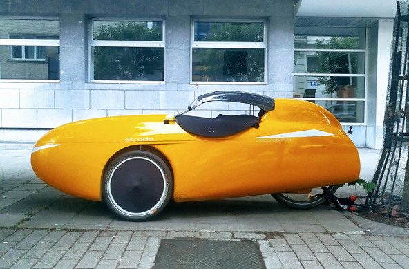 Fond ton gris bleu, mur d'un bâtiment dont on voit les fenêtres.
Devant se mur, le sol, trottoir gris bleu.
Sur ce trottoir, un vélo très particulier en forme de voiture de course jaune orangée.
Il s'agit d'un tricycle. 