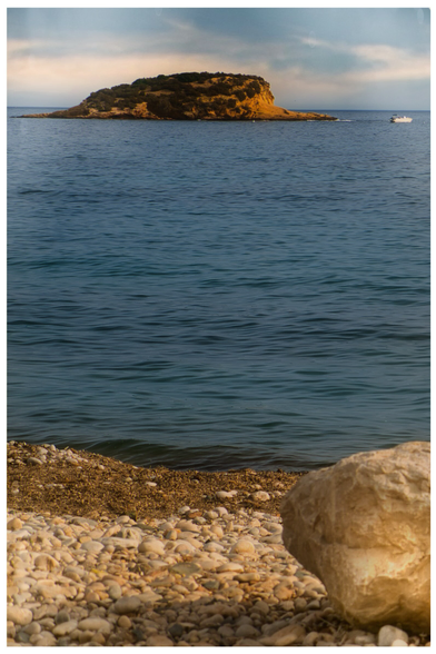 Hochformat: Im Vordergrund Meeresufer,  rechts ein großer Stein,  blaugrünes sanftes Meer , eine Felseninsel mit spärlichem Bewuchs, ein kleines weisses Boot, dunstiger bewölkter Himmel