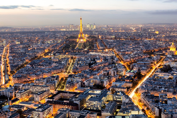 The image depicts a cityscape during dusk or early evening, characterized by a dense array of buildings and streets illuminated by streetlights and building lights. The city's layout features wide boulevards and a grid-like pattern. Prominently visible in the center is the Eiffel Tower, indicating that the city is Paris. The surrounding area is densely populated with buildings, and the city extends into the distance with numerous lights creating a vibrant, glowing effect. The sky is transitioning from day to night, with a gradient of colors from light to dark.