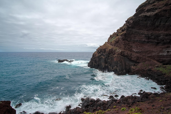 Fotografía en la q se ve el oceano en el centro y tercio inferior izquierdo chocando contra las rocas en un acantilado rocoso la parte izquierda del cuadro, en el tercio superior se aprecia el cielo gris y nuboso, algunas manchas de vegetacion dan un toque de color verde a la imagen.