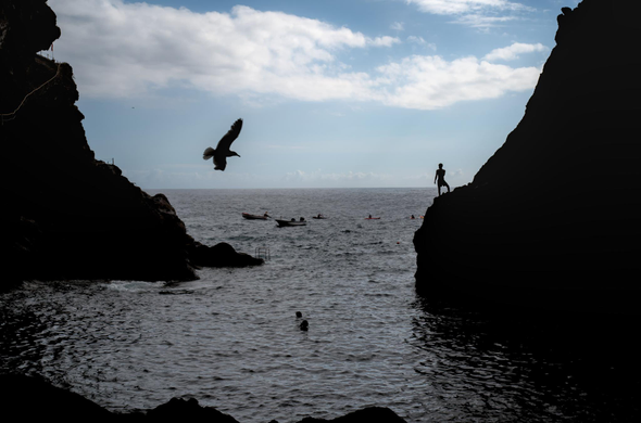 Fotografía desde el interior de una cueva al nivel del mar donde se ven las silietas de una gaviota a la izquierda y un chico subido a una piedra a la derecha, en el centro del cuadro se ven a dos personas dentro del agua y unas barcas en la lejania, tambien se ve el cielo azul y nubes