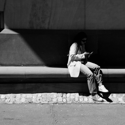 A black and white photo shows a woman sitting alone on a stone ledge against a large building wall. She is looking down at her smartphone, with both hands holding it. The shadow from the building casts a triangular shape across the wall and ledge, creating a strong contrast of light and dark.