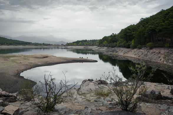 Una fotografia de un embalse en la q aparecen en primer plano en el tercio inferior dos arbustos en un suelo rocoso, una pasa de agua ocupa la parte central de la imagen donde se refleja el cielo y el bosque que se ve entrando en la imagen desde el a lado derecho, en el centro de la imagen hay una lengua de tierra q entra en el agua y en el extremo de esa lengua hay 3 personas, 1 de ellas pescando, tambien se reflejan en el agua, en la parte superior en la lejania se ve la presa y montañas q se pierden entre las nubes grises.