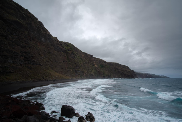 Fotografía de un acantilado donde se ve a la derecha el oceano con olas surcandolo una playa negra a la izquierda y el acantilado, en un día nuboso y gris.