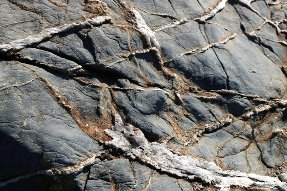 Close up photograph of the surface of a rock formation near a river. The rock is dark grey, with brown and white lines of sparkly crystal like veins on the surface.