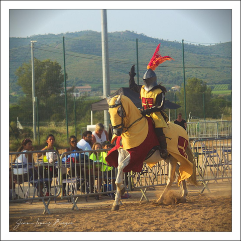A mounted knight in medieval-style armor and costume, riding a white horse, is in a sandy arena. The knight is wearing a dark gray/black helmet with reddish-orange plumes. The knight's armor and cape are a mix of yellow and maroon/reddish tones. 
The horse is wearing a matching, yellow and gold looking cloth over its neck. The ground is light tan/beige sand. A metal fence with various heights of metal fencing lines the perimeter of the arena. Several people are seated behind the metal fencing, watching the performance. Green hills and a partly cloudy sky are visible in the background.  A light beige/tan colored tent is visible in the background.