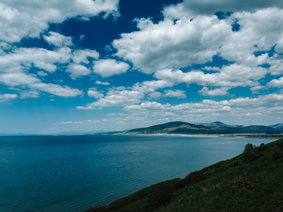 Landscape photo showing the sea, some hills in the distance, and the town of Barmouth. The weather is sunny but partly cloudy.