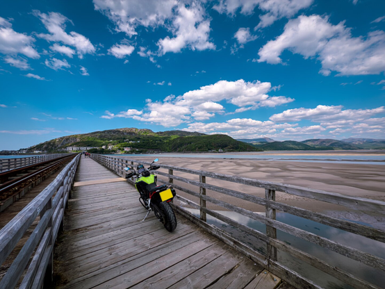 A motorcycle parked on a wooden bridge, next to a railway line, which crosses a tidal estuary. In the distance is a hill and the town of Barmouth.