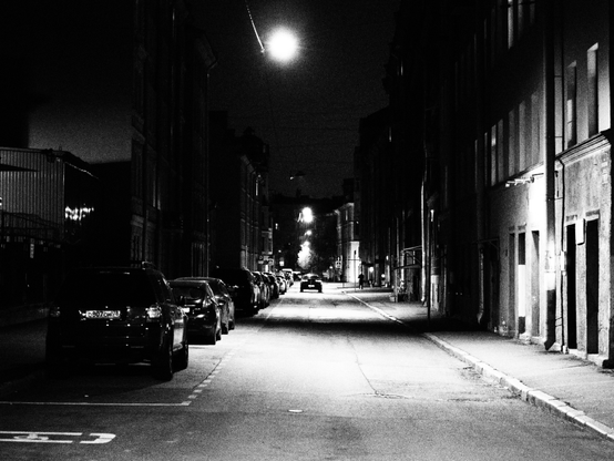 The late evening photo of the typical Saint-Petersburg small street in the center of the city. The street is lit by lamps, hanged on the wire above the street. On the left there are a parking slots with a lot of cars in it. On the right there are living houses built in the end of XIX / start of XX centuries.
Photo is black and white.